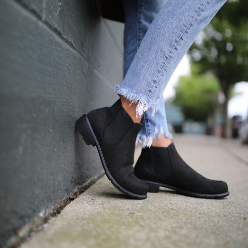 A person wearing light blue, frayed-edge skinny jeans and black ankle boots leans against a dark green wall on a city sidewalk, with trees and buildings blurred in the background.